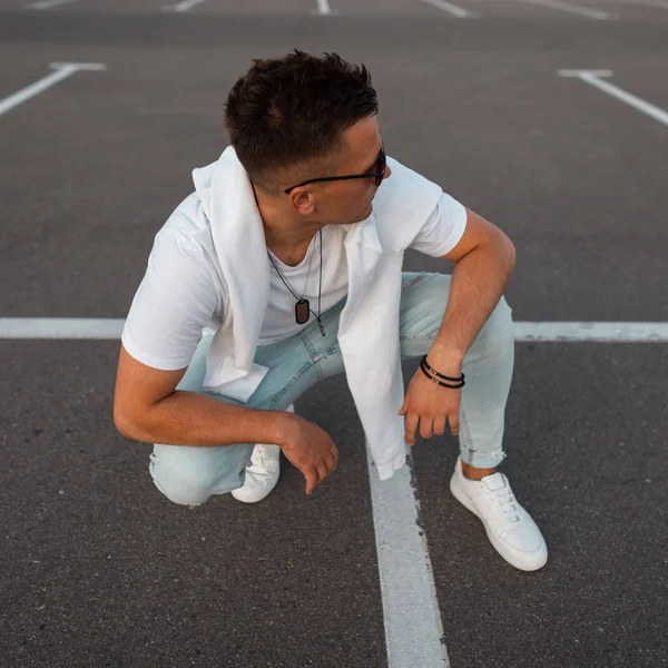 Homem hipster jovem na moda em jeans elegantes em uma camiseta branca vintage em óculos de sol da moda com uma camisola sentar-se no estacionamento ao ar livre. Cara atraente descansando em uma estrada com linhas brancas . — Fotografia de Stock