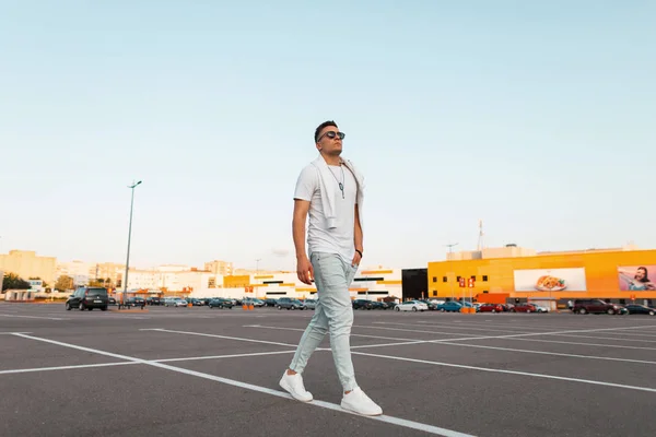 American Young Man hipster in zonnebrillen in modieuze kleding in trendy witte sneakers reist door de straat. Knappe stedelijke kerel in de moderne parkeerplaats in de stad. Stijlvolle jeugd zomer herenkleding — Stockfoto