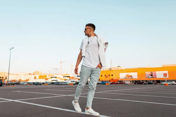 Amerikaanse jonge hipster man in zonnebril in Vintage blauwe jeans in een stijlvol wit T-shirt in Sneakers wandelingen door de stad. Stedelijke kerel wandelen in de straat. Zomer jeugd mode. Modieuze herenkleding — Stockfoto