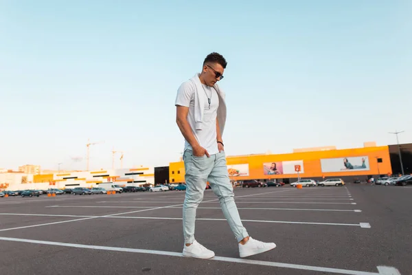 Homem hipster jovem na moda em jeans elegantes em uma camiseta branca vintage em óculos de sol na moda com uma camisola caminha ao longo de uma linha branca no asfalto. Um tipo urbano moderno viaja pela rua. Roupa masculina . — Fotografia de Stock