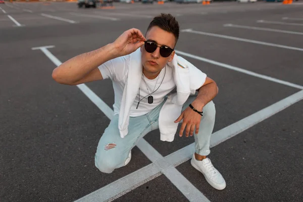 Jeune homme hipster beau en lunettes de soleil en jeans bleus à la mode dans un élégant T-shirt blanc en baskets est assis sur la route en ville un jour d'été. Un type qui se repose dans la rue. Mode de rue été — Photo