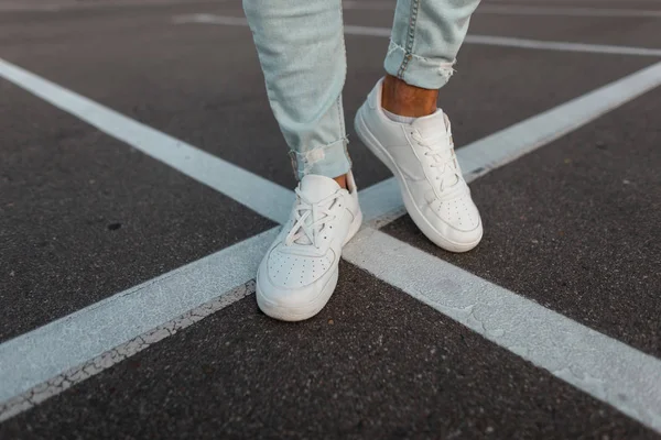 Close-up van mannelijke benen. Modieuze jonge man staat op de asfalt weg in lederen stijlvolle witte sneakers in trendy blauwe jeans. Seizoensgebonden heren schoenen. Straat casual stijl. — Stockfoto