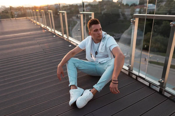 Vintage spor ayakkabı bir t-shirt şık bir saç modeli ile Avrupalı genç adam gün batımında bir cam duvar yakınında katta oturan dinleniyor. Modern adam bir yaz açık oyun üzerinde dinleniyor.