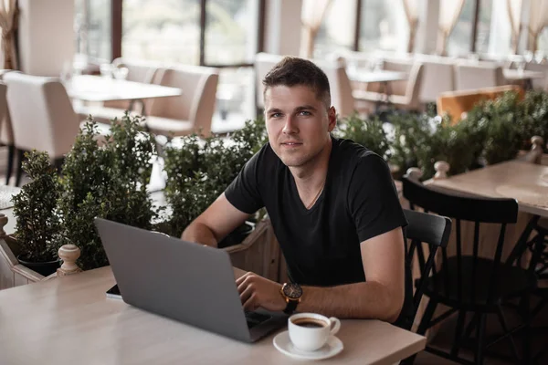 Úspěšný šťastný mladý obchodní muž v černém tričku s moderním počítačem sedí ve staré kavárně. Radostný chlápek na volné noze pracuje na přenosném počítači. Pracovní doba. — Stock fotografie