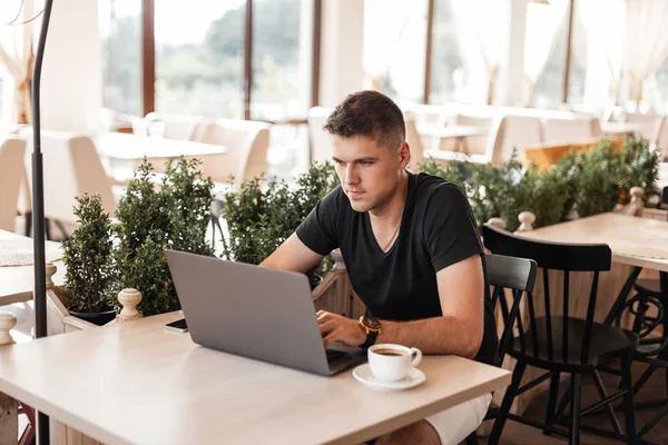 Pohledný obchodní mladík v elegantním tričku s laptopem sedí v kavárně a psát na klávesnici. Profesionální Blogger pracuje na internetu. Vzdálená práce. — Stock fotografie