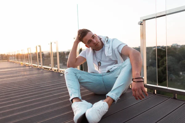 Çekici genç erkek modeli şık saç modeli açık düzlük. Şık giysiler yakışıklı trendy adam gün batımında açık bir terasta ahşap bir zemin üzerinde oturan dinleniyor.