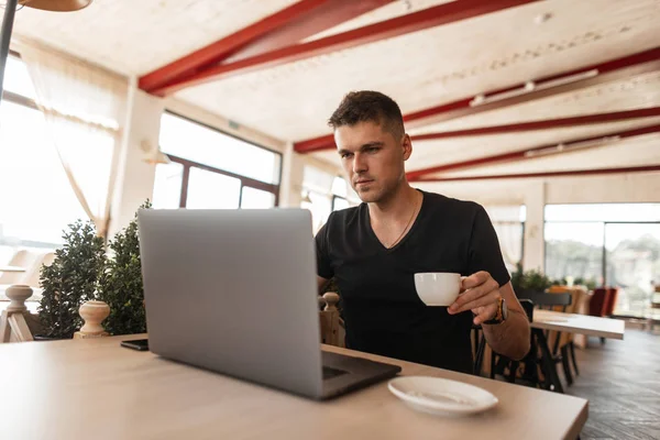 Mladý pohledný obchodník v černém tričku sedí ve staré kavárně a pracuje na moderním laptopu. Blogger pracuje online a pije kávu. Vzdálená práce. — Stock fotografie