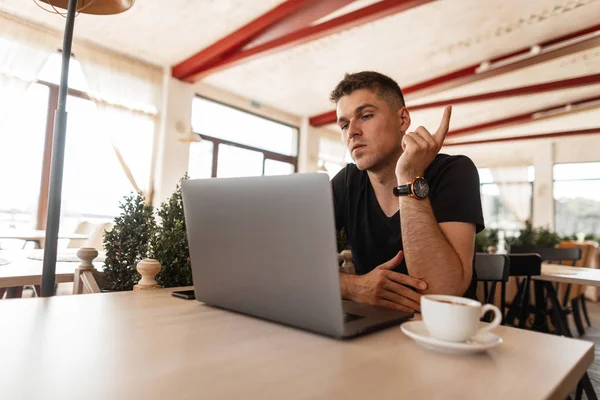 Mladý atraktivní obchodní muž ve stylovém černém tričku, který sedí v kavárně a pracuje vzdáleně na moderním notebooku. Pohledný chlápek na volné noze. Pracovní doba. — Stock fotografie