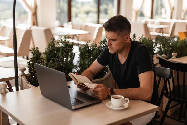 Pohledný mladý student v módním černém tričku s notebookem s šálkem kávy v kavárně si čte knihu. Atraktivní chlápek na volné noze rád čte v poledne. Koncepce vzdělávání. — Stock fotografie