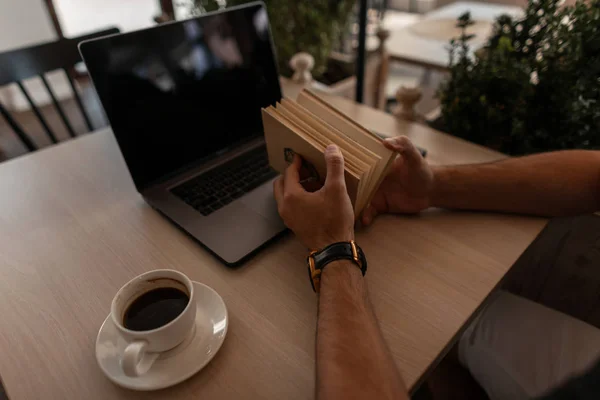 Blíží se k vám knížka s knihou. Mladý muž sedí v kavárně s notebookem s šálkem kávy a čte zajímavou knihu. — Stock fotografie
