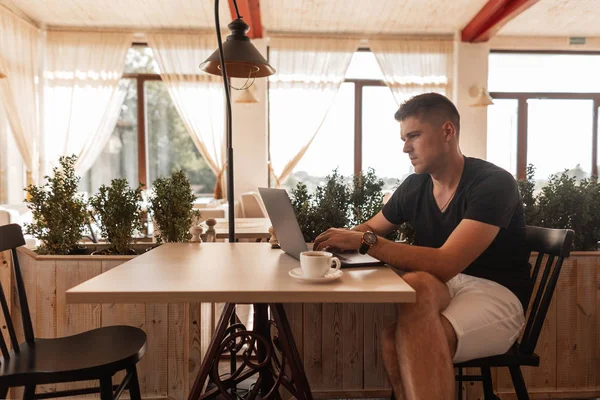 Şık genç adam şık giysiler içinde serbest bir vintage kafede bir masada oturur ve öğle yemeği saatinde modern bir dizüstü bilgisayar üzerinde çalışır. İş adamı. Uzaktan çalışma.