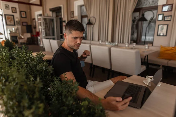 Şık bir t-shirt başarılı yakışıklı genç freelancer bir dizüstü bilgisayar ile vintage bir kafede ahşap bir masada oturur ve telefona bakar. Çekici blogger canlı online yayın yapıyor. 