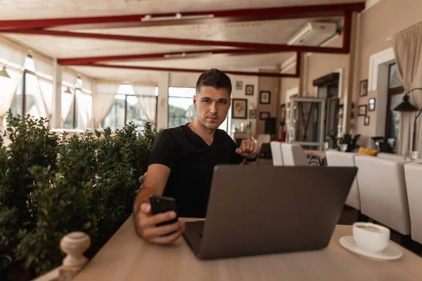 Úspěšný mladý blogger ve stylových šatech vysílá přes telefon živě z telefonu, zatímco sedí ve staré kavárně. Moderní obchodní chlapík se dívá do telefonu. Vzdálená práce. — Stock fotografie