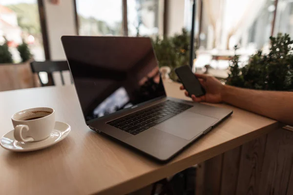 Blízko stolu, kde sedí obchodní muž s černým telefonem v ruce. Blízko pracoviště, na kterém je moderní laptop a šálek s kávou. Pracovní doba. — Stock fotografie