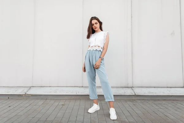 Hermosa joven morena con un elegante top de encaje en pantalones azules vintage en zapatillas blancas está de pie cerca de un edificio en la ciudad. Linda chica al aire libre. Colección de moda de verano de ropa de mujer Fotos De Stock Sin Royalties Gratis