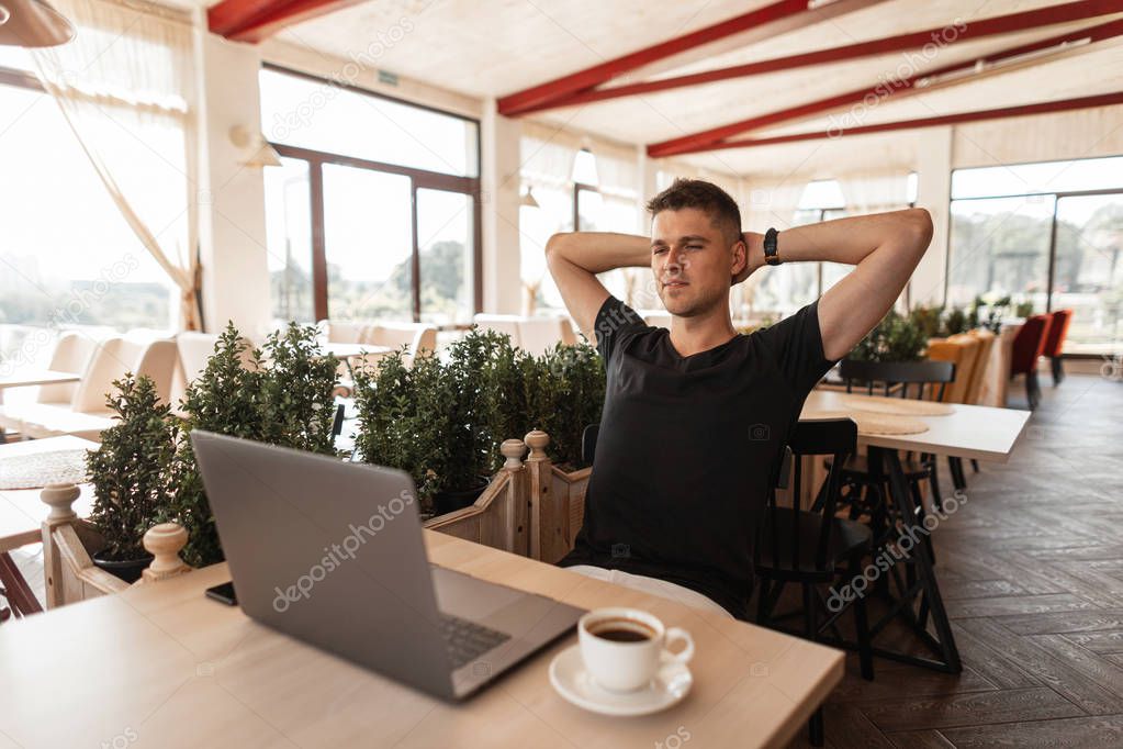 Successful joyful young business man in a black t-shirt with a computer sits in a modern cafe. Happy freelancer guy working remotely on a laptop.  Relax time.