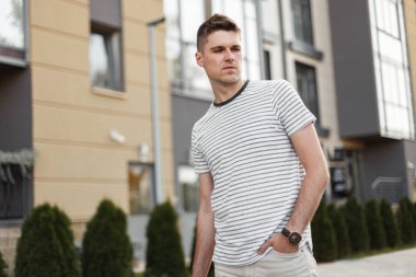 Bir saat ile şık bir t-shirt başarılı genç adam serbest bir yaz gününde modern bir binanın yakınında şehirde bir sokakta duruyor. Yakışıklı adam modeli bir yürüyüş sahiptir. Yaz trendy erkek giyim.