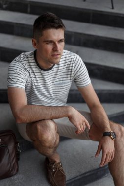 Şehirde bir taş merdiven üzerinde oturan işten sonra dinlenmiş bir deri vintage çanta ile şık süet kahverengi ayakkabı şık giysiler genç iş yakışıklı adam. Çekici adam dinlenme nin tadını çıkarıyor.