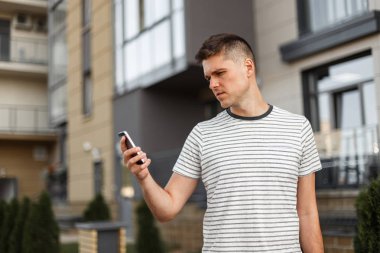 Şık bir saç modeli ile şık bir çizgili t-shirt genç yakışıklı adam yöneticisi şehirde modern bir binanın yanında elinde bir cep telefonu ile duruyor. İş adamı dışarıda. Modern teknoloji