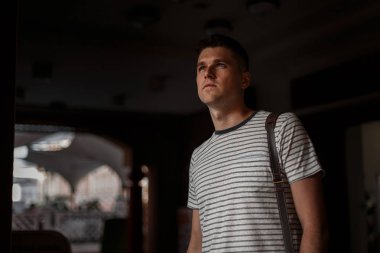 Omuz bir deri çanta ile trendy çizgili t-shirt şık bir saç modeli ile Avrupa güzel genç adam karanlık bir odada duruyor ve yukarı bakıyor. Stüdyodaki yakışıklı adam. Moda erkek giyim.