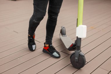 Spor ayakkabılı kot pantolon şık adam bir yaz gününde şehir etrafında bir elektrikli scooter sürmek. Erkek bacaklarının ve modern scooter'ın yakın çekim. Modern teknolojiler. Aktif yaşam tarzı.