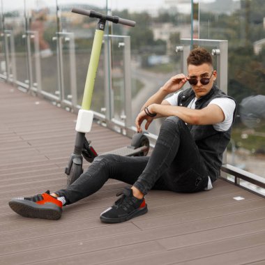 Güneş gözlüğü moda denim siyah giysiler serin amerikan hipster adam şehirde bir yaz kafenin ahşap zemin üzerinde oturup. Yakışıklı adam bir elektrikli scooter sürme sonra açık havada rahatlatır.