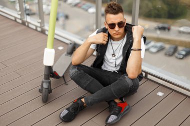 Bir cam duvar a yanında bir elektrikli scooter yanında ahşap bir zemin üzerinde oturan spor ayakkabı şık gri kot bir kot yelek güneş gözlüğü genç modern hipster adam. Adam şehirde hafta sonu sahiptir.