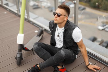 Bir elektrikli scooter bir yolculuk tan sonra yaz terasında ahşap bir zemin üzerinde güneş gözlüğü spor ayakkabı bir vintage kot yelek yakışıklı genç hipster adam. Şık adam açık havada rahatlatır