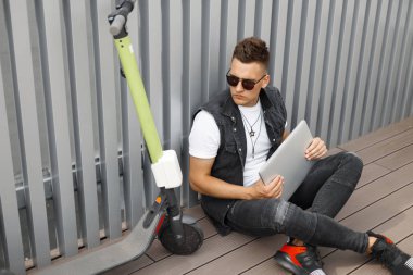 Şık güneş gözlüğü şık siyah denim giysiler yakışıklı genç adam hipster bir elektrikli scooter yakınında modern bir dizüstü bilgisayar ile katta oturur. Trendy Amerikalı adam metal bir duvarın yanında dinleniyor.