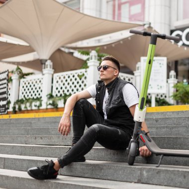 Güneş gözlüğü trendy siyah kot giysiler çekici genç adam hipster bir yaz kafe yakınında vintage taş adımlar üzerinde bir elektrikli scooter yanında oturur. Yakışıklı adam bir scooter sürdükten sonra rahatlatıcı sahiptir.