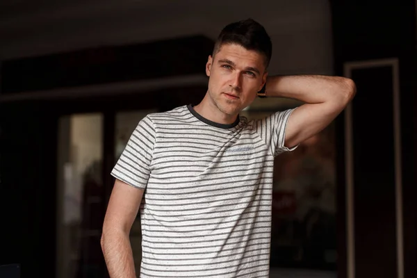 Modelo joven guapo con peinado elegante con hermosos ojos grises en una camiseta a rayas de moda posando de pie en una habitación oscura. Un tipo atractivo. Ropa de verano de moda para hombres . — Foto de Stock