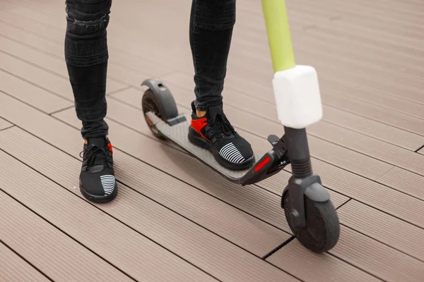 Close-up de pernas de homem em uma scooter elétrica moderna. Cara elegante em jeans em tênis fica em uma scooter na cidade em um dia de verão . — Fotografia de Stock