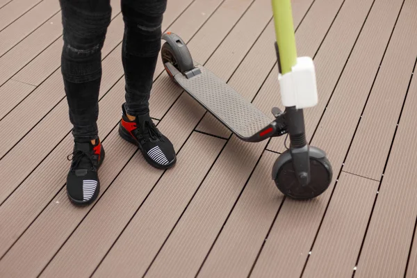 Männerbeine in grauen Jeans in trendigen schwarzen Turnschuhen neben einem Elektroroller an einem Sommertag. Der modische Typ steht auf einem Holzboden in der Nähe eines modernen Motorrollers. Nahaufnahme. — Stockfoto