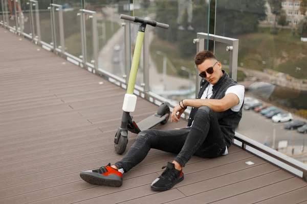 Young urban guy in fashionable gray jeans clothes in trendy black sunglasses sits next to an electric scooter on a wooden floor near a glass wall. Attractive guy enjoys relaxes after riding a scooter.