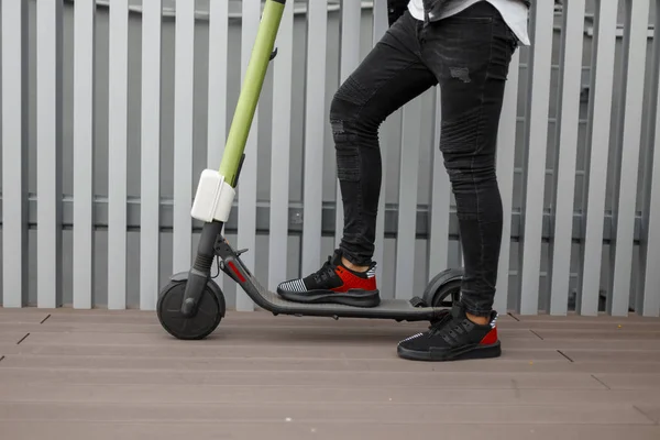 Nahaufnahme von Männerbeinen auf einem modernen Elektroroller. Stylischer Typ in Jeans und Turnschuhen steht an einem Sommertag auf einem Motorroller in der Stadt nahe der metallic-grauen Wand. — Stockfoto