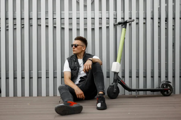 Élégant jeune homme hipster en lunettes de soleil sombres dans des vêtements d'été à la mode reposant après avoir roulé sur un scooter électrique près d'un mur métallique dans la ville. Américain mec est relaxant sur la terrasse en plein air . — Photo