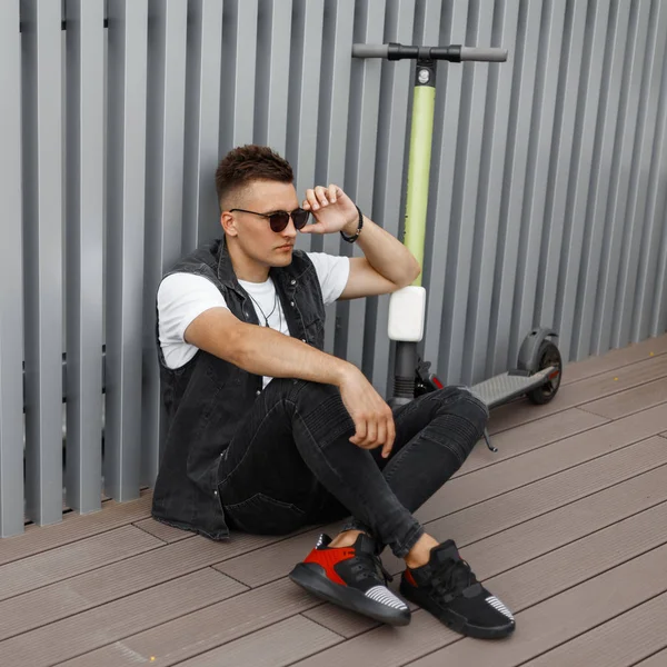 Модель молодого хіпстера людини в сонцезахисних окулярів в стильному одязі джинсів з скутером сидить на терасі біля металевої стіни. Гай сидить на підлозі поруч з електричний скутер. Релаксації. — стокове фото