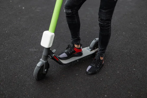 Eleganti gambe maschili in jeans neri in scarpe da ginnastica nere alla moda su uno scooter elettrico in una giornata estiva. Ragazzo alla moda si trova su un asfalto vicino a uno scooter moderno. Primo piano . — Foto Stock