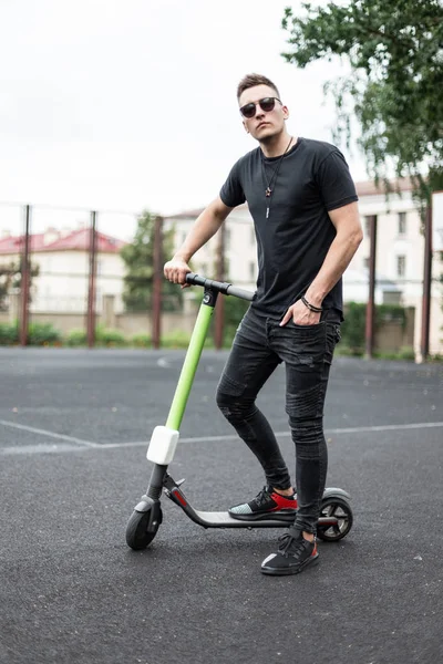 Jeune homme séduisant hipster en T-shirt noir en jeans en baskets en lunettes de soleil se tient avec un scooter électrique moderne sur un terrain de basket-ball en plein air. Guy se détend après une balade active sur un scooter . — Photo