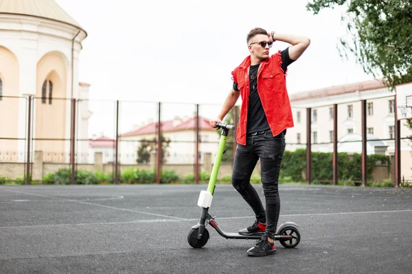 Giovane hipster in sneakers in un elegante gilet di denim rosso in jeans neri in occhiali da sole stand con uno scooter elettrico su un campo da basket in città. Ragazzo figo in posa con uno scooter all'aperto . — Foto Stock