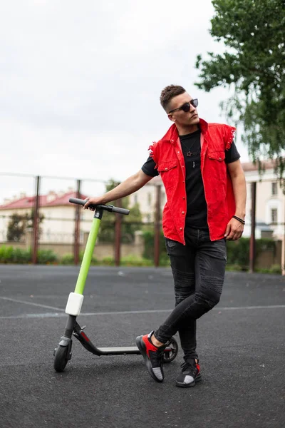 Attraente modello giovane uomo con uno scooter elettrico vintage in denim alla moda vestiti estate nero-rosso in occhiali da sole si rilassa sul campo da basket. Guy sta riposando dopo aver guidato uno scooter moderno . — Foto Stock