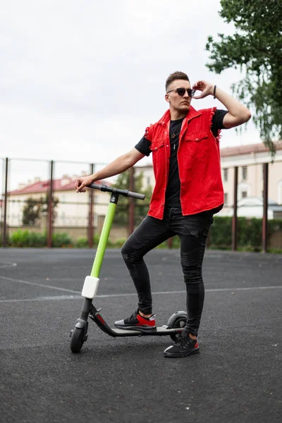 L'hipster giovane con un'acconciatura elegante in abiti di denim estivi alla moda è dotato di uno scooter vintage e raddrizza gli occhiali da sole. ragazzo americano cavalca un moderno scooter elettrico su un campo da basket . — Foto Stock