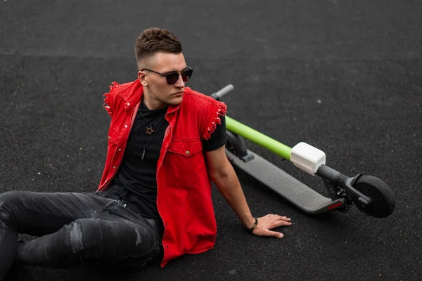 Handsome young hipster man with a hairstyle in stylish denim red-black clothes in sunglasses sitting on a basketball court next to a modern electric scooter. American cool guy is relaxes outdoors.