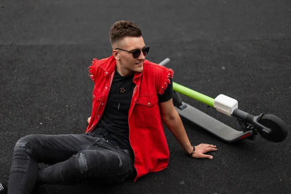 Positive cute young man in fashionable clothes in sunglasses sits and smiles near an electric scooter on the street. Happy hipster guy relaxes on a basketball court after riding on a modern scooter.