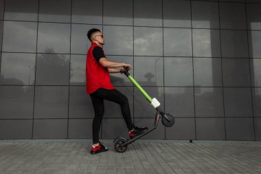 Siyah bir binanın yakınında modern bir elektrikli scooter ile poz güneş gözlüğü şık denim kırmızı-siyah giysiler Içinde Kentsel genç hipster adam. Modern serin adam bir yaz günü şehir etrafında bir scooter sürme.