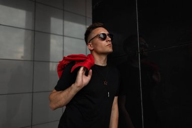 Siyah bir t-shirt kırmızı şık kot yelek ile şık güneş gözlüğü güzel bir genç adam modeli şehirde siyah bir duvar yakınında duruyor. Havalı bir hipster. Erkek giyim trendy yaz koleksiyonu.