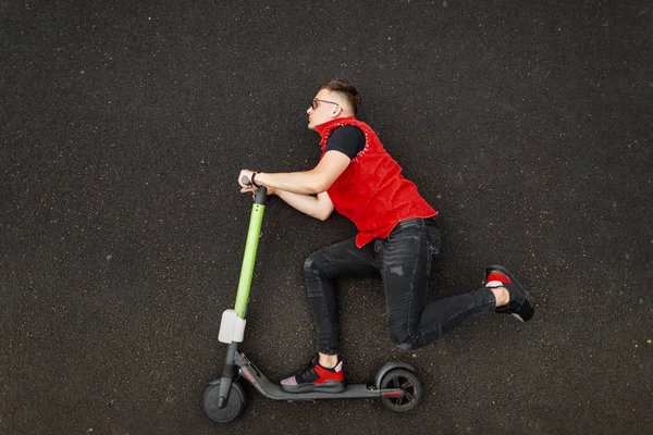Attrayant jeune homme hipster dans un gilet à la mode en jeans dans des chaussures de gym en lunettes de soleil posant couché avec un scooter sur l'asphalte dans la ville. Un beau mec en scooter. Vêtements pour hommes. Vue d'en haut . — Photo