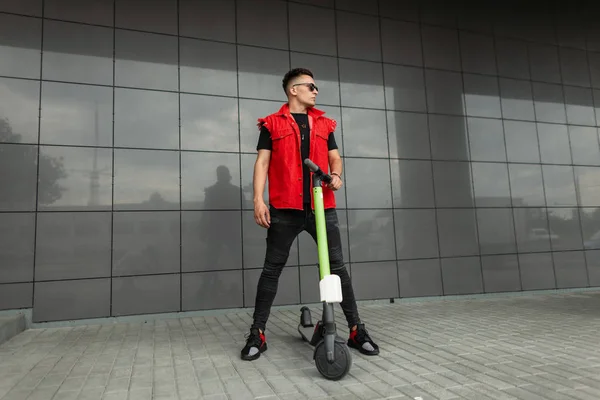 Joven hombre fresco hipster posando con scooter eléctrico. El tipo con ropa de vaqueros negros rojos de moda en gafas de sol con un scooter está descansando de pie cerca de una pared moderna oscura en la ciudad. Estilo de calle . — Foto de Stock