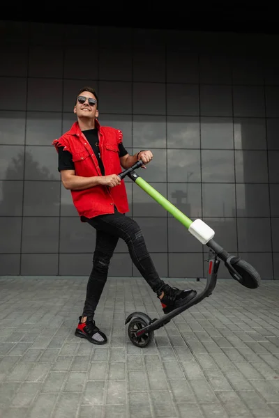 Gioioso giovane hipster con taglio di capelli in eleganti abiti denim rosso-nero in occhiali da sole posa con un moderno scooter elettrico vicino a un edificio grigio. Allegro ragazzo fresco cavalcando uno scooter in giro per la città . — Foto Stock