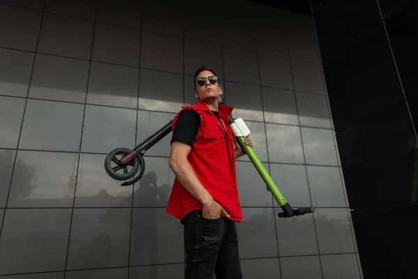 Young cool man hipster posing with electric scooter. Guy in fashionable red- black jeans clothes in sunglasses with a scooter on his shoulder is resting standing near a dark modern wall in the city.